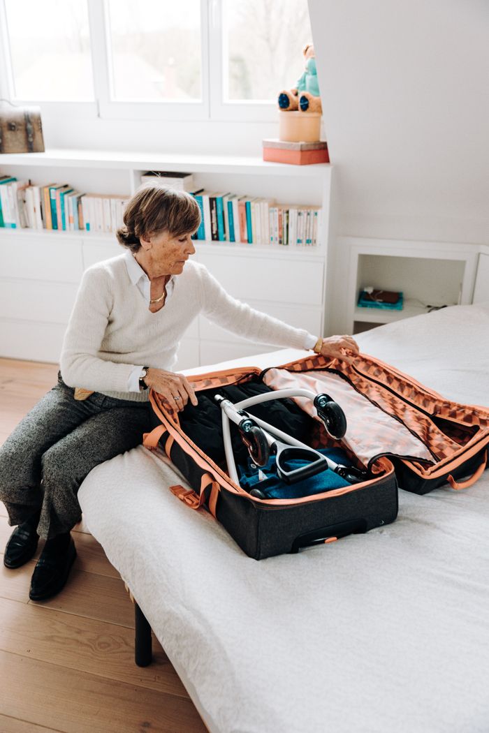 Senior woman carefully packs her rollator into a suitcase, preparing for travel while sitting on a bed.